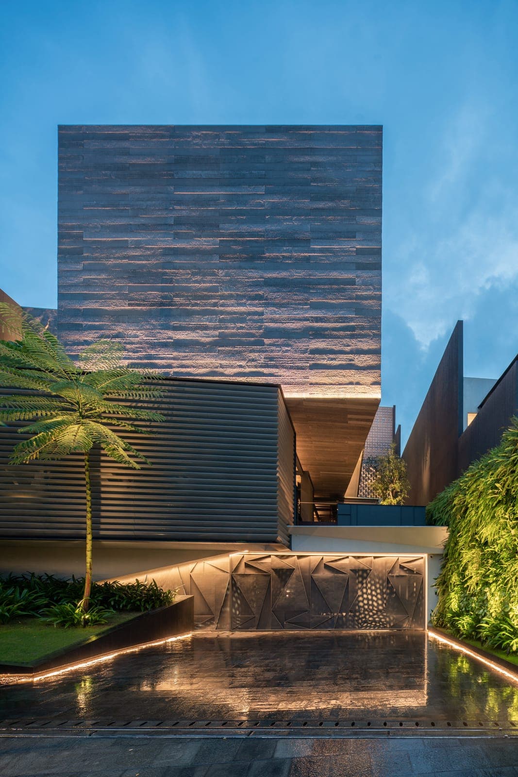 The entry at dusk: banded stone raised over louvres, and a faceted panel lit above water.